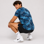 A man crouching in a studio setting, wearing a blue camouflage short-sleeve athletic t-shirt, black shorts, white Nike socks, and white Nike Air Max sneakers against a light gray background.