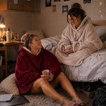 Two women laughing in a cozy bedroom, each wrapped in a plush hooded blanket and holding a mug. One wears a deep burgundy blanket, and the other wears a light beige blanket.