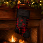 A Christmas stocking with a red and black plaid pattern and embroidered red snowflakes, hanging in front of a warm fireplace with a garland above.