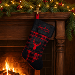 A personalized Christmas stocking with a red and black buffalo plaid pattern, a dark gray velvet cuff embroidered with the name 'Archie', and a red embroidered deer head patch, hanging on a wooden mantel decorated with a warm fireplace and holiday garland.