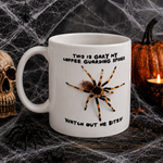A white ceramic Halloween mug with a printed spider and the text 'THIS IS GARY MY COFFEE GUARDING SPIDER WATCH OUT WE BITES!' surrounded by spooky props including a skull, pumpkins, and fake cobwebs.