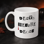A white ceramic mug with the text 'DEATH BEFORE DECAF' printed in black block letters, featuring a small red drip detail. The mug is set on a dark, smoky surface with autumn leaves and a small pumpkin in the background.