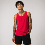 A man with visible tattoos wears a bright red tank top and black shorts against a plain light gray studio background.