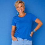 A smiling woman with short blonde hair wearing a bright blue crewneck t-shirt and light blue jeans, standing against a solid blue background.