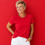 A smiling woman with short blonde hair wearing a bright red short-sleeve crewneck t-shirt and white pants, posing against a solid red background.