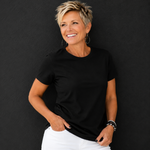 A woman with short blonde hair wearing a plain black short-sleeve crewneck t-shirt and white pants, posing against a dark textured wall.
