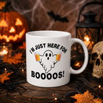 A white ceramic Halloween mug with the text 'I'M JUST HERE FOR BOOOOS!' and a ghost graphic holding orange drinks, styled on a rustic wooden surface with a jack-o'-lantern, skull, spider, lantern, and autumn leaves in the background.
