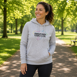 A woman wearing a light gray hoodie with a 'Kirkcaldy Parks Running Festival' graphic stands on a tree-lined path in a park.