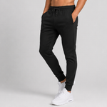 A person modelling black jogger sweatpants with a drawstring waist, side pockets, and ribbed ankle cuffs, paired with white sneakers against a light grey studio background.
