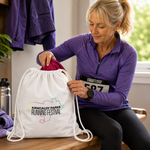 A woman in a purple running shirt sits on a bench and reaches into a white drawstring bag printed with the Kirkcaldy Parks Running Festival logo. The bag has white drawstrings and a pink interior lining.