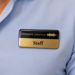 A rectangular staff badge with a black top half and gold bottom half, pinned to a light blue dress shirt. The top half reads 'DYNAMITE LIFESTYLE' with a gold soundwave graphic, and the bottom half reads 'Staff'.