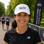 A smiling woman wearing a white Kirkcaldy Parks Running Festival cap and a black New Balance t-shirt, standing outdoors with race participants blurred in the background.