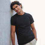 A man wearing a black short-sleeve crewneck t-shirt and light beige pants, leaning against a textured light gray concrete wall.