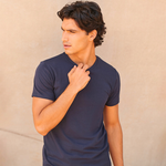 A man wearing a dark navy blue short-sleeve crewneck t-shirt, posing with his hand near his neck against a textured beige wall.