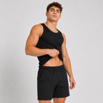 A muscular man wearing a black sleeveless tank top and black athletic shorts, lifting the hem of the tank top to show his midsection against a light gray studio background.
