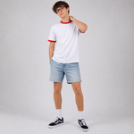 A young man wearing a white ringer t-shirt with red trim, light blue denim shorts, white socks, and black sneakers, posing against a light gray studio background.