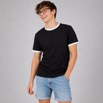 A man wearing a black short-sleeve ringer t-shirt with white trim on the collar and sleeve cuffs, paired with light blue denim shorts, standing against a solid gray studio background.