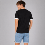 A man wearing a black ringer t-shirt with white trim on the collar and sleeve cuffs, paired with light blue denim shorts, photographed from the back against a solid gray studio background.