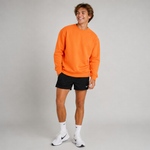 A man modeling a bright orange crewneck sweatshirt, black athletic shorts, white socks, and white sneakers against a light gray studio background.