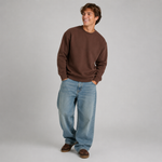 A man wearing a brown crewneck sweatshirt, light wash blue jeans, and brown shoes, standing against a light gray studio background.