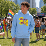 A young man standing on a grassy outdoor crowd at a pride event, wearing a light blue hoodie with a yellow and purple 'BUT CHILL GAY' graphic and light-wash jeans.