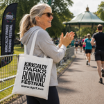 A woman at a park running event carries a white tote bag printed with 'KIRKCALDY PARKS RUNNING FESTIVAL' and the website.