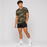 A man modeling a short-sleeve crewneck t-shirt with an olive green and brown camouflage pattern, paired with black athletic shorts, white socks, and white sneakers, set against a plain light gray studio background.
