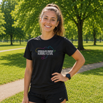 A smiling woman stands outdoors on a park path wearing a black short-sleeve athletic T-shirt with a colorful 'Kirkcaldy Parks Running Festival' graphic on the chest, paired with black shorts.
