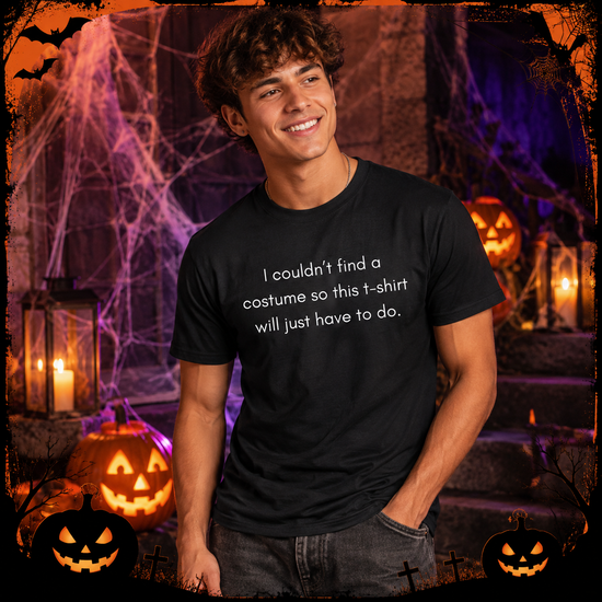 A smiling young man wearing a black graphic t-shirt with white text that reads 'I couldn't find a costume so this t-shirt will just have to do.' He is posed in a Halloween-themed setting with jack-o'-lanterns, cobwebs, and candles.