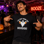 A man wearing a black graphic t-shirt with a white ghost and beer mug design that reads 'I'M JUST HERE FOR BOOOOS!' points at the shirt in a lively, dimly lit bar or party setting with a neon 'BOOZE' sign in the background.