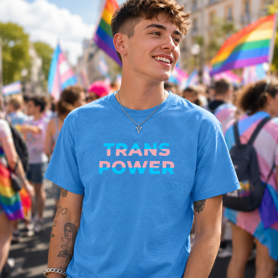 A smiling young man at a pride parade wearing a bright blue t-shirt with 'TRANS POWER' printed in pink and blue gradient text across the chest. The background is filled with blurred rainbow and transgender pride flags.