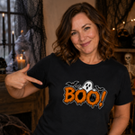 A woman wearing a black Halloween t-shirt with a 'BOO!' graphic, pointing to the design. The background is a cozy, decorated Halloween room with candles, spiderwebs, and pumpkins.