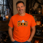 A smiling man wearing a bright orange graphic T-shirt with a Halloween 'BOO!' design featuring bats, a ghost, and a jack-o'-lantern, posed in a cozy, decorated Halloween-themed room.