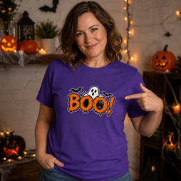 A woman wearing a purple Halloween t-shirt with a 'BOO!' graphic, pointing to the chest print in a cozy, decorated Halloween-themed room.