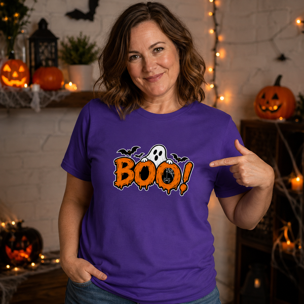 A woman wearing a purple Halloween t-shirt with a 'BOO!' graphic, pointing to the chest print in a cozy, decorated Halloween-themed room.