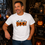 A man wearing a white graphic t-shirt with a Halloween 'BOO!' design, posed in a cozy, decorated Halloween room with pumpkins, a skeleton, and candles.