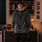 A man wearing a dark grey oversized fleece hoodie, black sweatpants, and black-and-white slide sandals stands in a dorm room setting with a bed, desk, and bookshelf in the background.