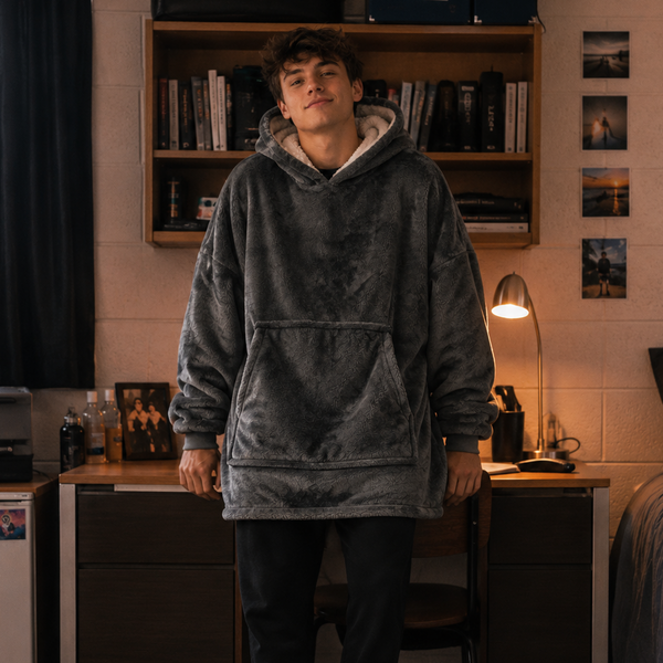 A man wearing a dark grey oversized fleece hoodie, black sweatpants, and black-and-white slide sandals stands in a dorm room setting with a bed, desk, and bookshelf in the background.