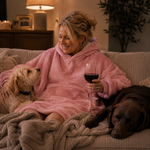 A woman relaxes on a couch wearing a plush pink hooded blanket robe while holding a glass of red wine, with two dogs beside her in a cozy living room setting.