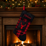 A personalized Christmas stocking with a red and black plaid pattern, a dark navy cuff and toe, and red embroidery featuring the name 'Harlow' and a deer head silhouette. The stocking is hanging from a gold hook on a rustic wooden mantel decorated with a lit Christmas tree garland and a warm fireplace in the background.
