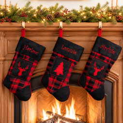 Three personalized Christmas stockings with red and black plaid patterns hang from a wooden fireplace mantel decorated with a green pine garland and warm string lights. The center stocking is embroidered with the name 'Lonnie' and features a red Christmas tree motif, while the side stockings feature red deer head motifs and the names 'Harlow' and 'Archie'.