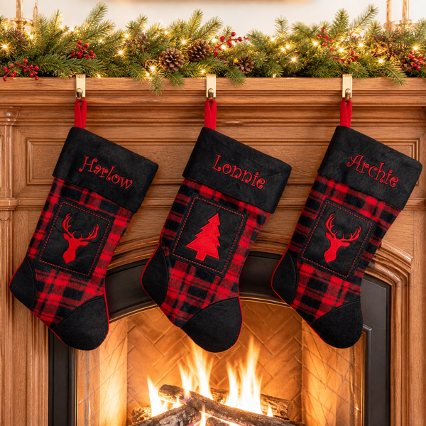Three personalized Christmas stockings with red and black plaid patterns hang from a wooden fireplace mantel decorated with a green pine garland and warm string lights. The center stocking is embroidered with the name 'Lonnie' and features a red Christmas tree motif, while the side stockings feature red deer head motifs and the names 'Harlow' and 'Archie'.