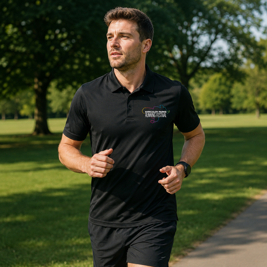 A man jogging outdoors in a black short-sleeve polo shirt and black shorts, with a green park and tree-lined path in the background. The polo shirt has a small white and yellow logo on the left chest