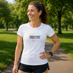 A woman wearing a white graphic t-shirt and black running shorts stands on a paved path in a sunny park with green trees in the background. The t-shirt features a colourful outline of Kentucky and the text 'KIRKCALDY PARKS RUNNING FESTIVAL'.