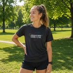 A woman standing in a sunny park wearing a charcoal short-sleeve t-shirt with a colourful 'Kirkcaldy Parks Running Festival' graphic on the chest, paired with black running shorts.