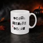 A white ceramic coffee mug with the text 'DEATH BEFORE DECAF' printed in black, featuring a small red blood drop detail. The mug is set on a dark, smoky surface with autumn leaves and a small pumpkin in the background.