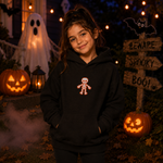 A young girl wearing a black hoodie with a small red and white skeleton graphic on the chest, standing in a spooky Halloween yard decorated with jack-o'-lanterns, a ghost, and a 'Beware Spooky Boo!' sign.