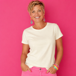 A smiling woman with short blonde hair wearing a plain cream short-sleeve t-shirt and bright pink pants, standing against a solid bright pink background.