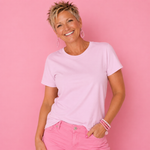 A smiling woman with short blonde hair wearing a light pink crew-neck t-shirt and pink pants, posing against a solid pink background.