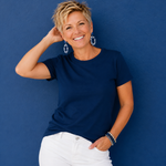 A smiling woman with short blonde hair wearing a navy blue short-sleeve crewneck t-shirt and white pants, posing against a textured blue wall.